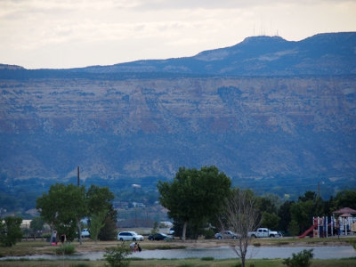 [A small portion of the early evening sky is above the canyon wall and is topped by at least six different antenna. In the foreground are the trees and cars at the park which are dwarfed by the canyon wall.]